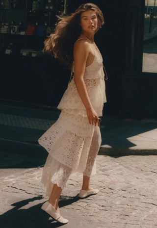 a-woman-wearing-a-white-lace-tiered-dress-and-white-flats-walks-on-a-cobblestone-street-in-warm-sunlight
