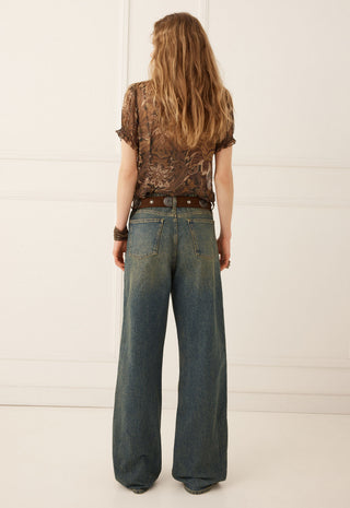 a-woman-viewed-from-behind-wearing-a-brown-patterned-short-sleeve-top-a-brown-belt-and-wide-leg-blue-jeans-against-a-white-paneled-wall