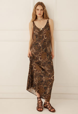 a-woman-modeling-a-sleeveless-brown-maxi-dress-with-a-metallic-bronze-floral-pattern-paired-with-brown-strappy-sandals-standing-against-a-white-paneled-wall