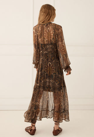 a-woman-viewed-from-behind-wearing-a-sheer-brown-patterned-midi-dress-with-long-sleeves-and-ruffled-cuffs-paired-with-brown-strappy-sandals-set-against-a-white-paneled-wall