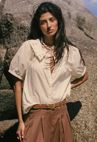 a-woman-standing-outdoors-against-a-rocky-cliff-wearing-a-cream-short-sleeve-blouse-with-a-ruffled-collar-and-buttons-brown-pleated-pants-a-brown-belt-and-layered-necklaces