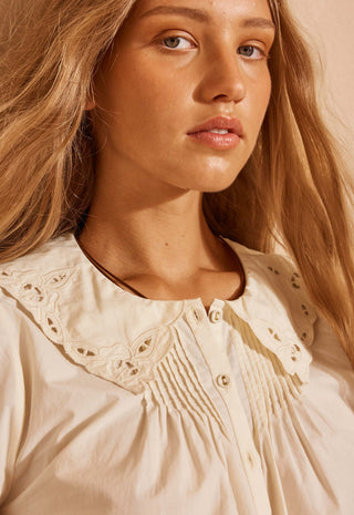 close-up-of-a-woman-wearing-a-cream-colored-blouse-with-a-large-eyelet-lace-collar-and-smocked-chest-detailing-set-against-a-warm-beige-background