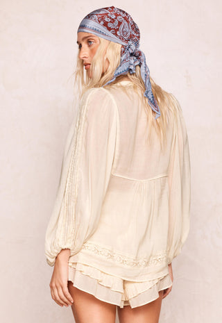 a-woman-shown-from-the-back-wearing-a-cream-colored-semi-sheer-long-sleeve-blouse-with-lace-trim-and-a-paisley-bandana-headscarf-set-against-a-textured-beige-wall