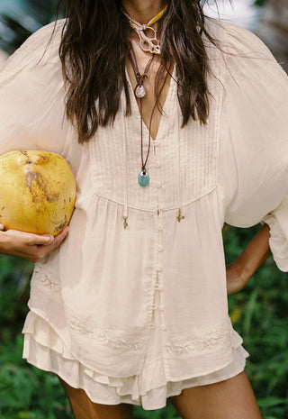 a-woman-wearing-a-cream-colored-bohemian-style-short-dress-or-romper-with-ruffled-edges-and-button-details-holding-a-yellow-coconut-in-her-hand-against-a-green-outdoor-background