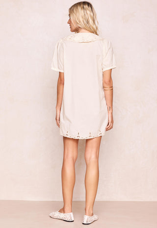 a-woman-shown-from-the-back-wearing-a-short-sleeve-cream-colored-tunic-dress-with-a-ruffled-collar-and-eyelet-lace-trim-at-the-hem-paired-with-white-lace-up-flats-against-a-textured-beige-wall