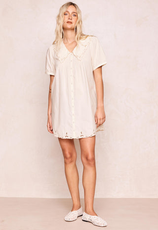 a-woman-modeling-a-short-sleeve-cream-colored-mini-dress-with-a-ruffled-peter-pan-collar-and-eyelet-lace-trim-paired-with-white-lace-up-flats-against-a-textured-beige-wall