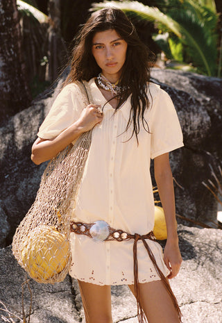 a-woman-stands-outdoors-on-rocky-ground-wearing-a-cream-short-sleeve-button-front-dress-or-tunic-with-a-fringed-hem-accessorized-with-a-brown-woven-belt-layered-necklaces-and-a-large-woven-straw-tote-bag