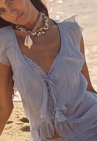 a-woman-on-a-sandy-beach-wearing-a-light-blue-ruffled-cover-up-top-with-lace-trim-and-a-shell-necklace