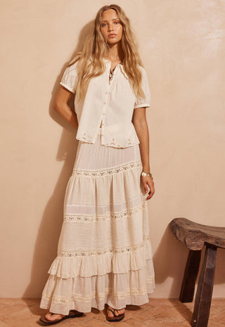 a-woman-modeling-a-bohemian-style-two-piece-outfit-consisting-of-a-short-sleeve-cream-button-up-shirt-and-a-matching-long-tiered-maxi-skirt-with-lace-details-set-against-a-warm-beige-textured-wall