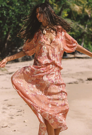 a-woman-wearing-a-flowing-peach-floral-maxi-dress-with-short-puff-sleeves-and-a-v-neckline-captured-mid-spin-on-a-sandy-beach-with-green-foliage-in-the-background