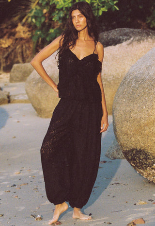 a-woman-stands-barefoot-on-a-sandy-beach-with-large-rocks-and-greenery-in-the-background-wearing-a-black-lace-camisole-and-matching-black-harem-style-pants