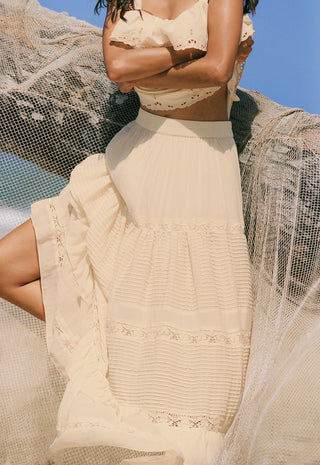 a-woman-wearing-a-cream-colored-tiered-maxi-dress-with-delicate-lace-and-embroidery-details-posed-outdoors-against-a-blue-sky-with-sheer-netting-draped-around-her