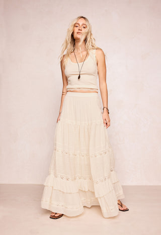 a-woman-modeling-a-cream-colored-two-piece-set-consisting-of-a-sleeveless-crop-top-and-a-long-tiered-maxi-skirt-with-lace-details-set-against-a-neutral-textured-beige-backdrop