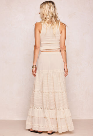 a-woman-shown-from-the-back-wearing-a-cream-colored-sleeveless-top-and-matching-tiered-maxi-skirt-with-lace-trim-standing-against-a-textured-beige-wall