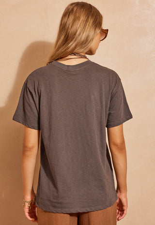 a-woman-viewed-from-behind-wearing-a-dark-charcoal-gray-short-sleeve-t-shirt-and-brown-pants-standing-against-a-textured-beige-wall