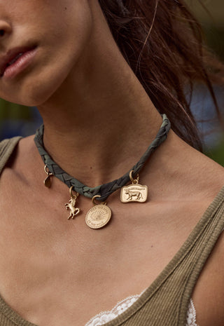 A close-up of a woman wearing a dark olive green braided leather choker necklace with gold-toned animal and circular pendant charms.