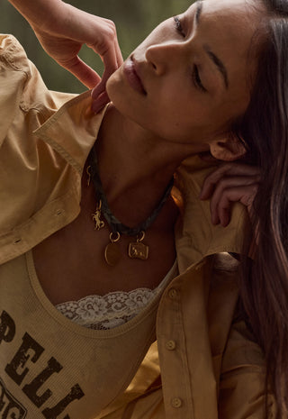 A close-up lifestyle shot of a woman wearing a layered necklace set. The set includes a dark green braided cord necklace with a small gold crescent moon and a gold square charm, layered over a gold chain with a gold 'N' initial pendant.