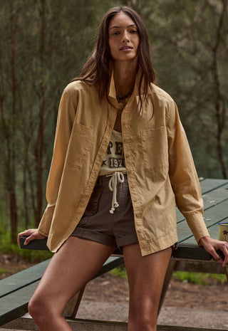 A woman wearing a tan long-sleeve button-up shirt over a graphic tee and brown shorts, leaning on a green picnic table in a wooded outdoor setting.