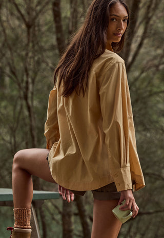 A woman wearing an oversized tan button-up shirt and brown shorts, posing outdoors in a wooded setting.