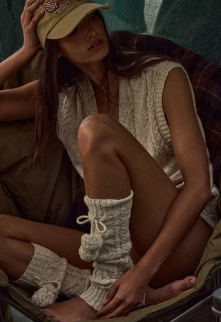 A woman seated in a camp chair wearing a beige knit sleeveless top, matching knit leg warmers with pom-poms, a tan baseball cap, and olive pants in a rustic outdoor setting.
