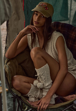 A woman sits in a camping chair wearing a beige knit sleeveless top, matching knit leg warmers, and a beige baseball cap with a red embroidered bee.
