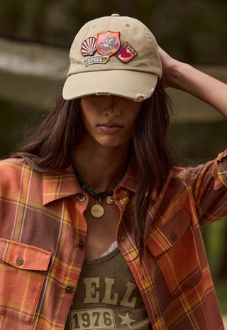 A woman wearing a beige baseball cap with multiple embroidered patches, an orange and brown plaid flannel shirt, and a graphic tank top, posed outdoors against a blurred green background.