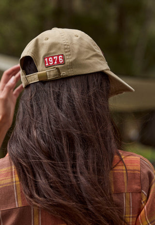 A person viewed from behind wearing a tan baseball cap with a red rectangular patch that reads 1976. The person has long dark brown hair and is wearing a brown and orange plaid shirt, with a blurred outdoor background.