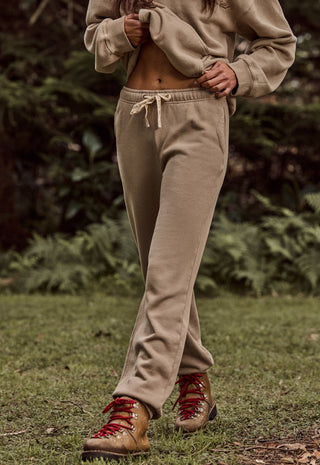 A person outdoors wearing loose beige jogger sweatpants with a drawstring waist, a matching beige cropped sweatshirt, and brown hiking boots with red laces, standing on grass with a blurred forest background.