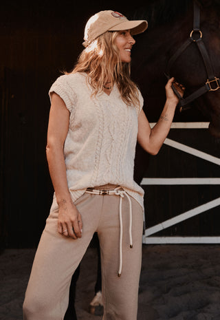A woman wearing a beige knit sleeveless top, matching beige drawstring pants, and a beige baseball cap stands in a barn next to a dark horse, holding its bridle reins.