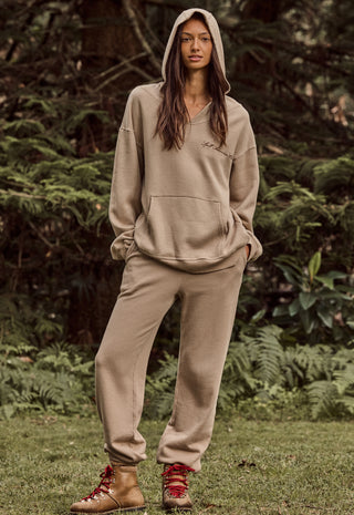 A woman stands outdoors in a wooded setting wearing a matching beige hooded sweatshirt and jogger pants set, with red lace-up ankle boots.