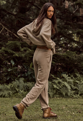 A woman wearing a beige cropped hoodie and matching jogger pants with brown boots, standing outdoors in a grassy area with a forest background.