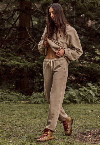 A woman wearing a matching beige cropped hoodie and sweatpants set with brown lace-up boots, standing in a grassy forest clearing.