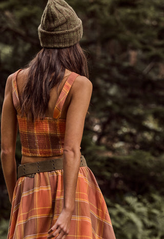 A woman viewed from behind wearing a sleeveless rust-orange plaid dress with a wide brown belt and a matching knit beanie, standing outdoors against a blurred forest background.
