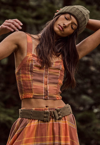 A woman wearing a rust-orange and brown striped knit top with a button front, a matching plaid skirt, and an olive green beanie, posed outdoors against a dark, blurred forest background.