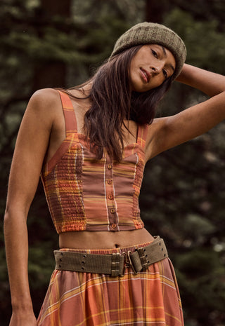 A woman wearing a rust-orange and brown plaid sleeveless crop top with button details, a matching plaid skirt, and a textured olive green beanie, photographed outdoors against a blurred forest background.