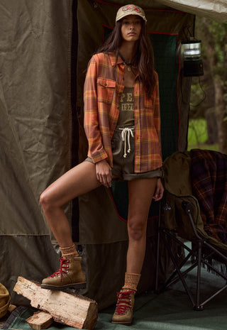 A woman posing in front of a camping tent wearing a rust-orange and brown plaid flannel shirt over a graphic tee