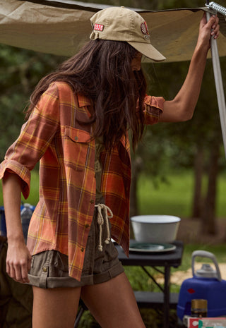 A woman wearing a rust-orange and brown plaid flannel shirt, olive shorts, and a khaki cap stands under a tent at a campsite.