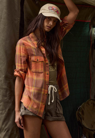 A woman models a rust-orange and yellow plaid button-up shirt with rolled sleeves, paired with olive shorts and a beige baseball cap, set against a dark tent background.