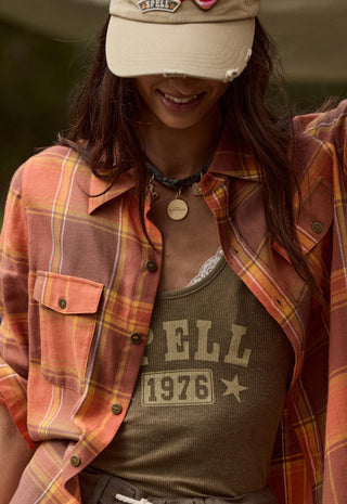 A woman wearing a rust-orange and yellow plaid flannel shirt over an olive green graphic tank top, paired with a beige baseball cap.