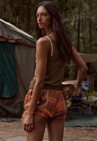 A woman standing outdoors in a campsite setting, wearing a sleeveless olive-brown top and rust-orange plaid shorts, with a tent and trees in the background.