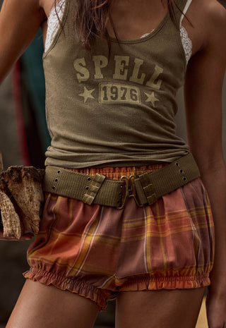 A close-up of a person wearing an olive green ribbed tank top with 'SPELL 1976' printed on the front, paired with rust-orange plaid bloomers and a wide olive belt.