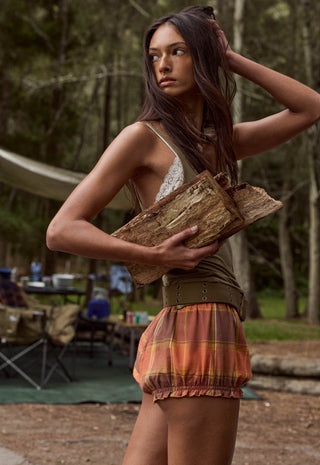 A woman standing outdoors in a wooded campsite setting, holding a piece of wood. She is wearing a sleeveless lace-trim top, an olive green belt, and a rust-orange plaid ruffled skirt.