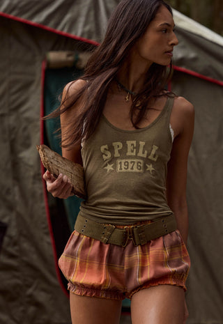 A woman wearing a vintage-style olive green tank top with 'SPELL 1976' printed on the front, paired with rust-orange plaid biker shorts and a matching belt, standing in front of a tent.