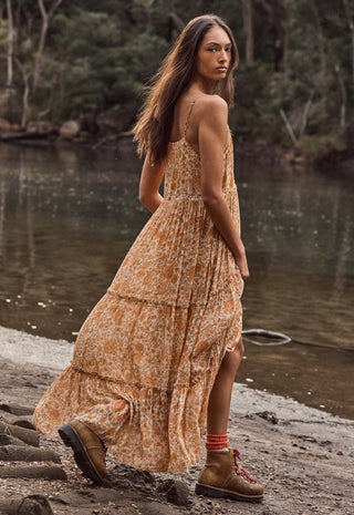 A woman wearing a flowing orange and beige floral maxi dress with brown hiking boots stands on a rocky riverside path with water and trees in the background.