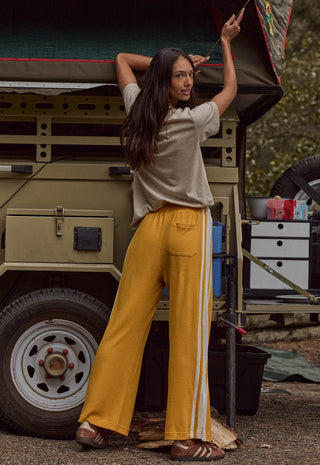 A woman stands outdoors beside a vintage military-style vehicle, wearing loose mustard yellow track pants with white side stripes, a light beige short-sleeve top, and brown sandals.