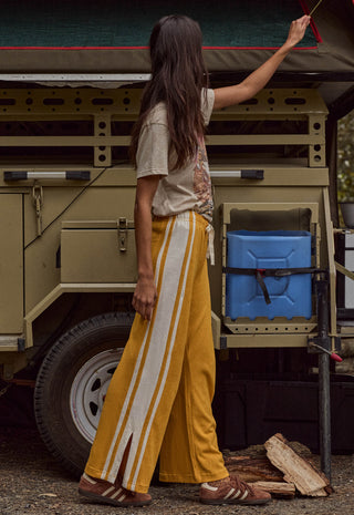 A woman stands in front of a vintage military-style vehicle wearing wide-leg mustard yellow pants with white side stripes, a beige graphic t-shirt, and brown sandals.
