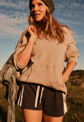 A woman outdoors wearing a beige oversized sweatshirt, dark brown shorts with white side stripes, and a dark knit beanie, with a camouflage jacket draped over her arm against a blue sky background.