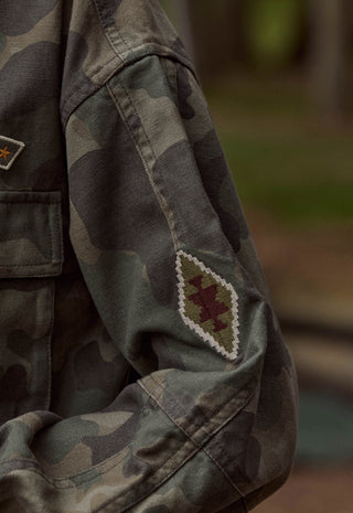 Close-up of a camouflage jacket sleeve with a diamond-shaped embroidered patch featuring a red star and white border, worn outdoors with a blurred background.