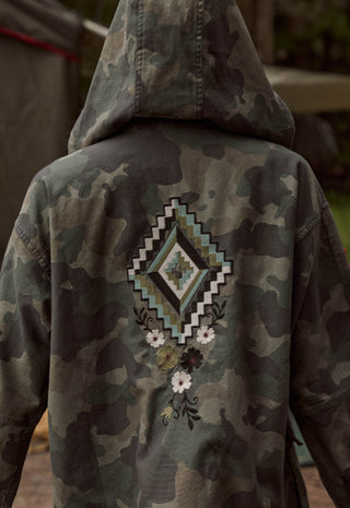 Back view of a camouflage hooded jacket with a large geometric and floral embroidered design on the back, shown on a person outdoors.