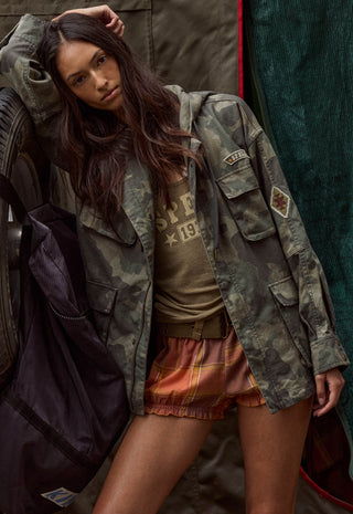 A woman posing in a vintage-style camouflage utility jacket over a graphic t-shirt and orange plaid shorts, leaning against a rustic background with a dark backpack.